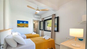 A hotel room with two beds, a ceiling fan, a painting, a window with curtains, and a bedside table with a lamp.