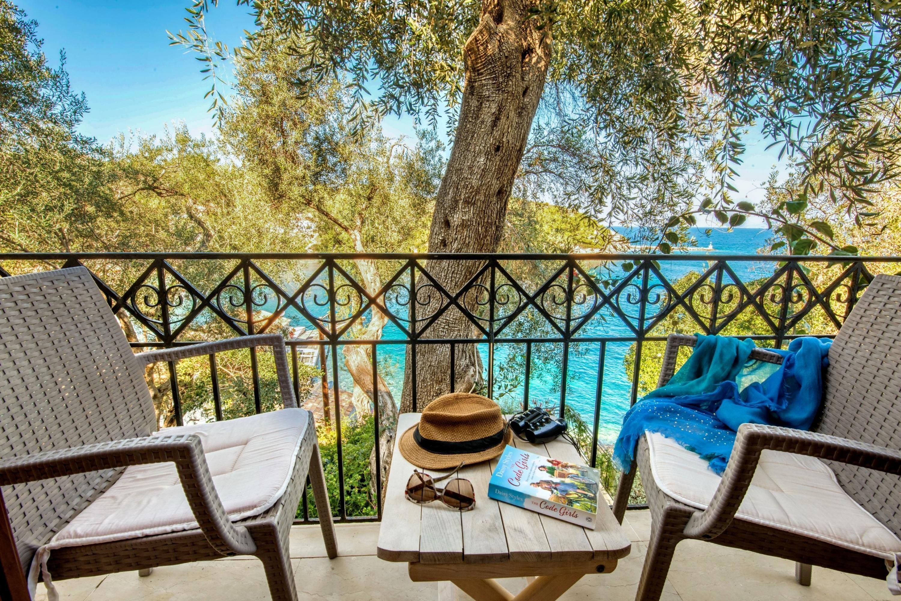 A balcony with wicker chairs, a table with a hat, sunglasses, and a book, overlooking a clear blue sea and lush greenery.