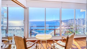 A room with a large window offering a view of the sea and a cityscape. There is a round table with chairs and a small table with a glass container and a glass. - Beach View 2D increible ubicacion (Valparaíso)