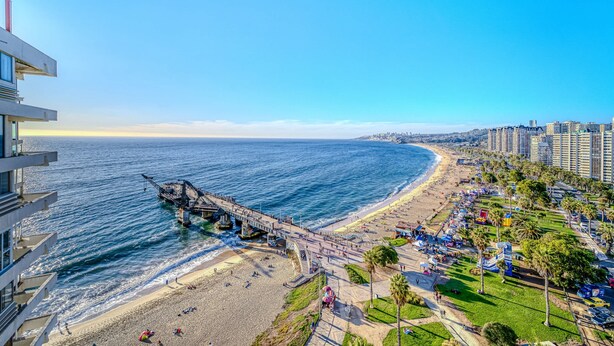 A coastal cityscape with a pier extending into the sea, a beach with people and umbrellas, and high-rise buildings in the background. - Beach View 2D increible ubicacion (Valparaíso)