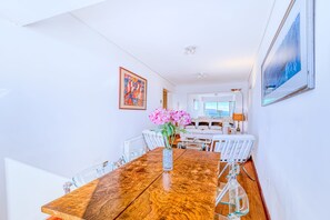 Un coin repas avec une table en bois, des chaises en verre transparent et un vase rempli de fleurs roses. Les murs sont blancs et un tableau encadré est accroché au mur.