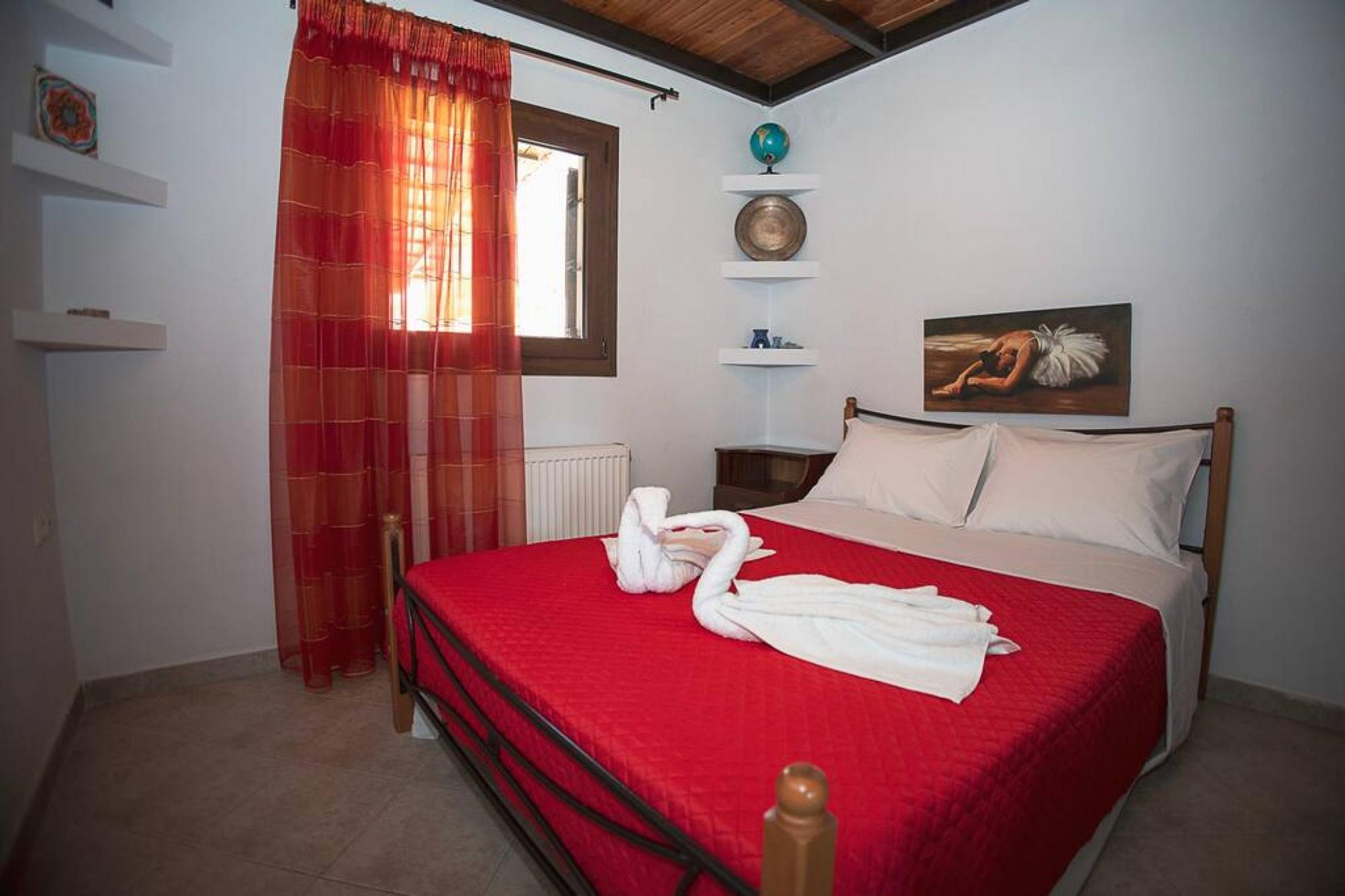 Een slaapkamer met een rood bedlaken, een houten hoofdbord, een wereldbol, een schilderij en een rood gordijn.