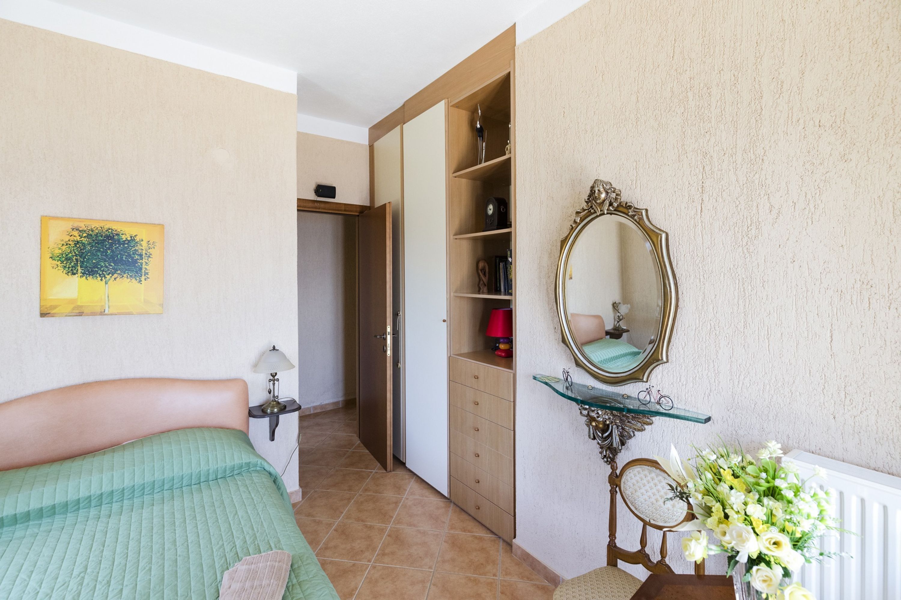 Un dormitorio con cama, armario, espejo, silla y un jarrón de flores.