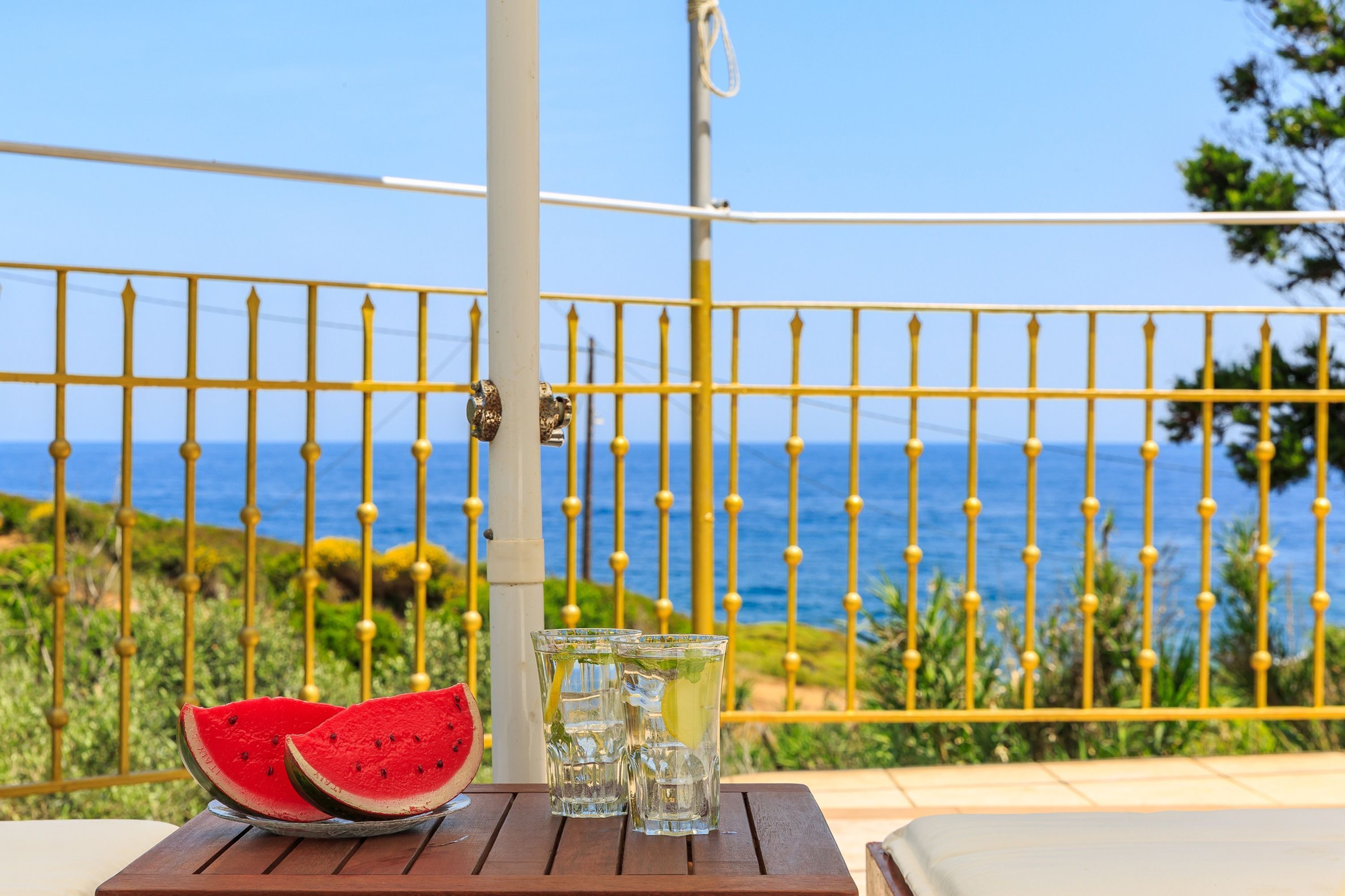 A wooden table with two glasses of water and a slice of watermelon on a balcony with a yellow railing and a view of the sea.