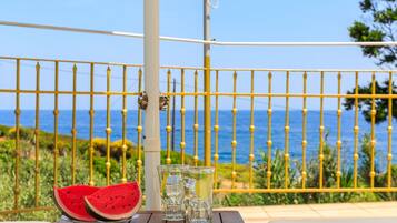 A wooden table with two glasses of water and a slice of watermelon on a balcony with a yellow railing and a view of the sea.