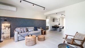 A modern living room with a sofa, two round wooden side tables, a chair, and a dining area in the background. - Two-Bedroom Apartment Sea View (Nikiana)