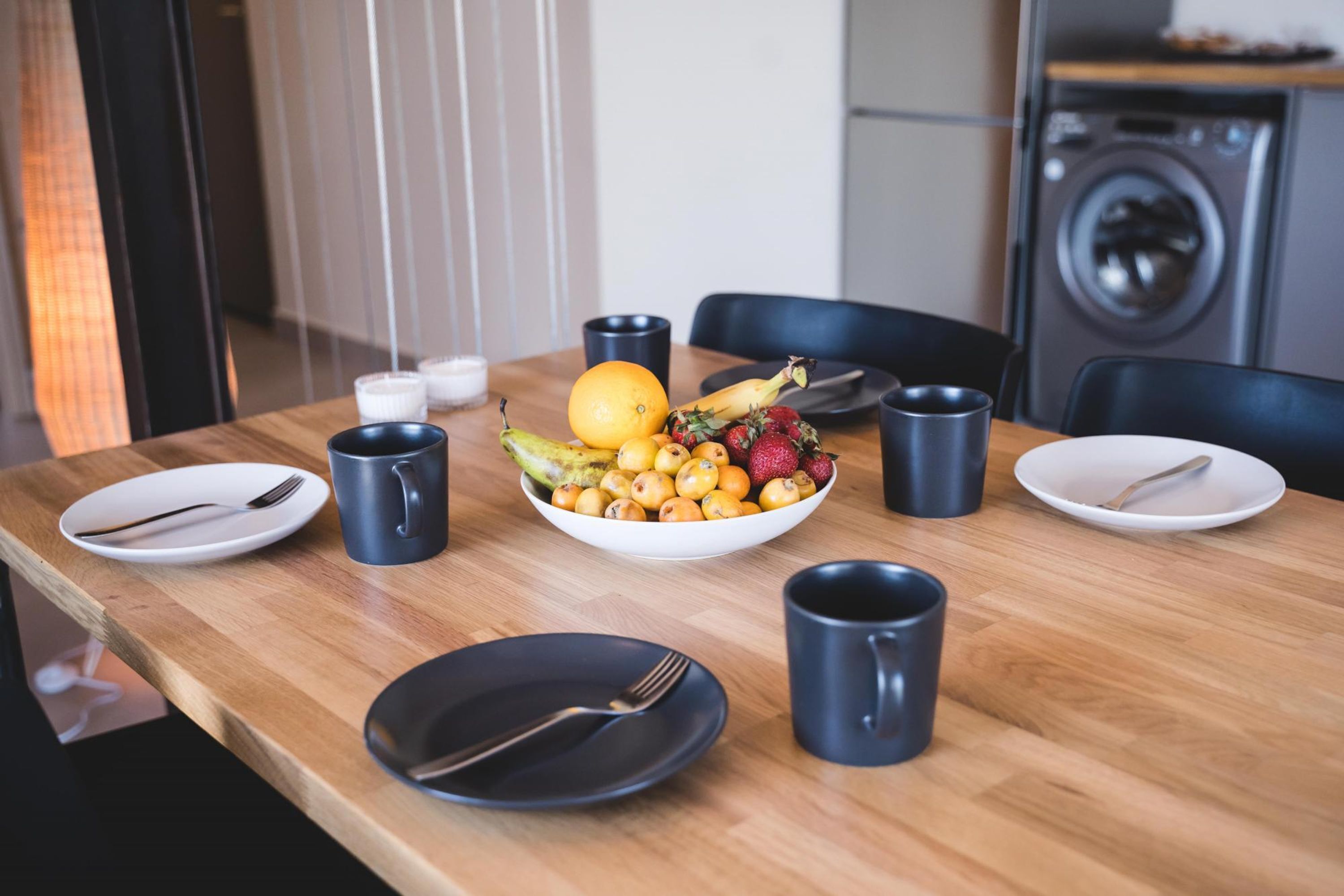 A dining table with a bowl of fruit, plates, cutlery, and black mugs.