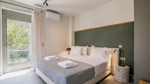 Quarto de hotel moderno com uma cama grande, mesa de cabeceira, luminária e vista para o verde através de uma porta deslizante.