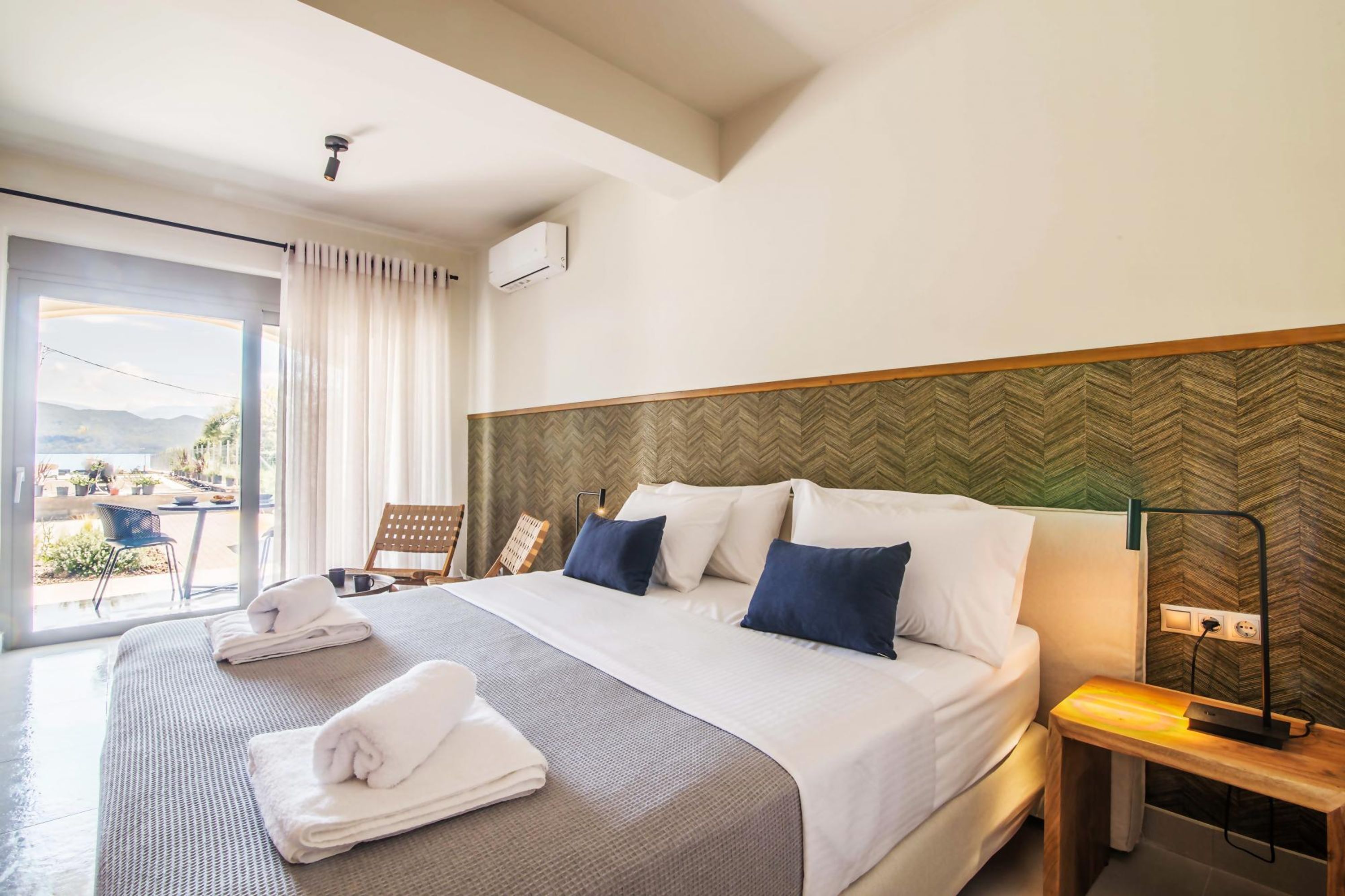 Een hotelkamer met een groot bed, een houten hoofdeinde, een nachtkastje met een lamp en uitzicht op het omliggende landschap.