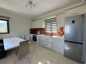 A modern kitchen with white cabinets, a stainless steel refrigerator, a dining table with chairs, and a window with blinds. - Suite 1 - Elpida Luxury Suites (Nydri)