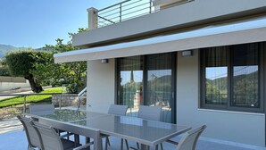 Un espace de restauration extérieur moderne, avec une table en verre et des chaises en métal, situé devant un bâtiment doté de grandes fenêtres et d’un balcon.