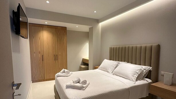 A modern bedroom with a large bed, a wooden wardrobe, a TV mounted on the wall, and a bedside table with a lamp. - Suite 5 - Elpida Luxury Suites (Nydri)