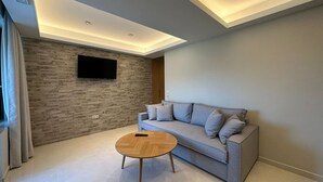 A modern living room with a grey sofa, a wooden coffee table, and a mounted TV on a textured wall. - Suite 5 - Elpida Luxury Suites (Nydri)
