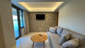 A modern living room with a sofa, a coffee table, a flat-screen TV mounted on a stone wall, and a large window with curtains. - Suite 5 - Elpida Luxury Suites (Nydri)