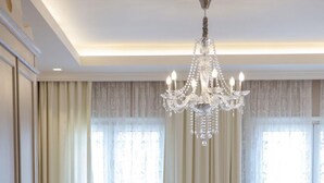 A spacious bedroom with a large bed, a chandelier, and a seating area with a table and chair. - Tirkouaz Suite - VK Luxury Suites-Private Parking (Nydri)