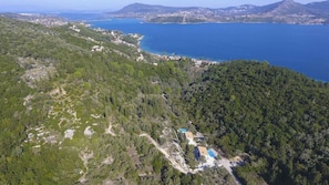 Luftaufnahme einer Küstenlandschaft mit dichtem Wald, einem kleinen Gebäude mit Pool und einer großen Wasserfläche.