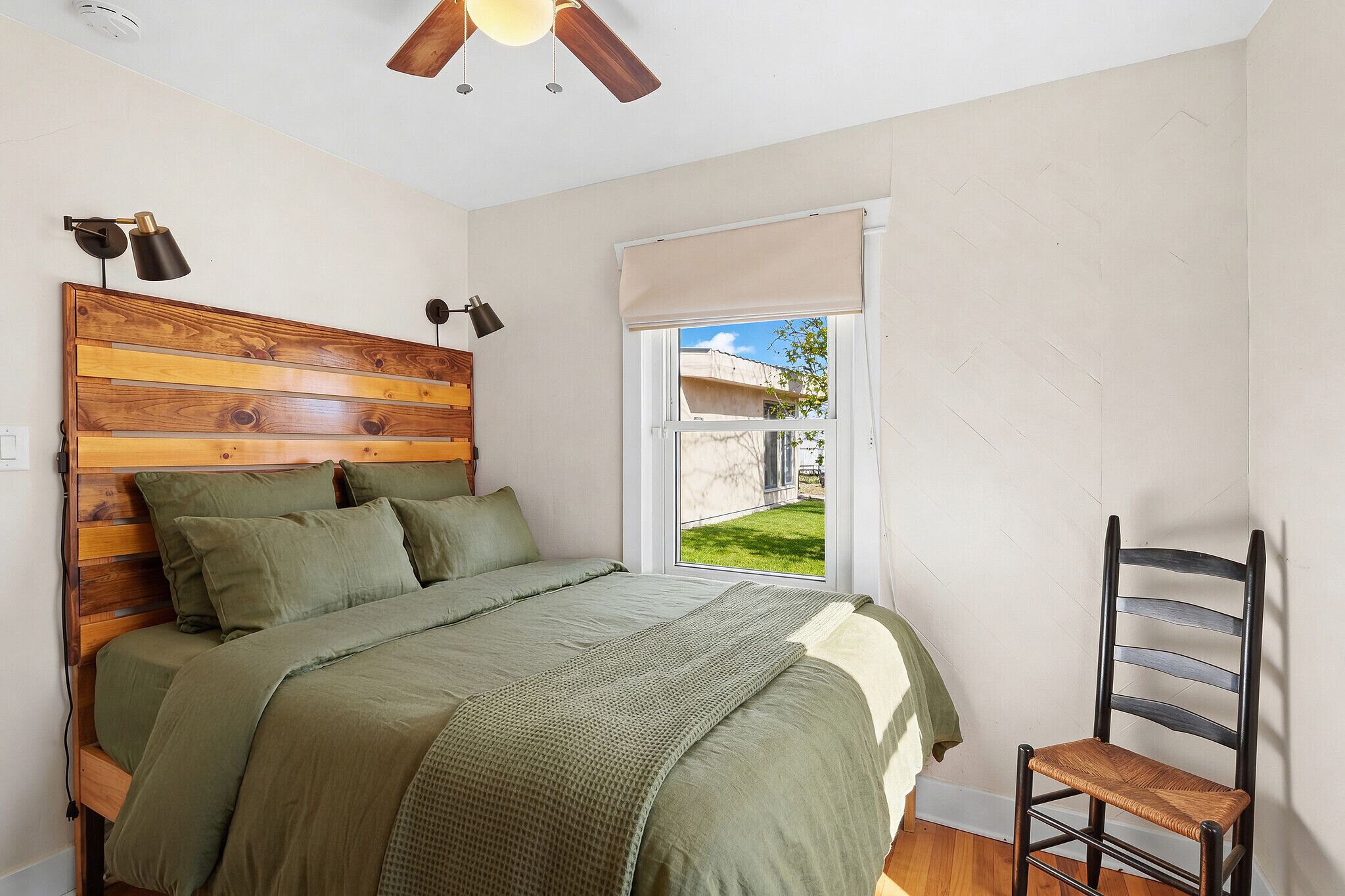 A bedroom with a wooden headboard, a bed with green bedding, a window with a view of the outside, a ceiling fan, and a chair.