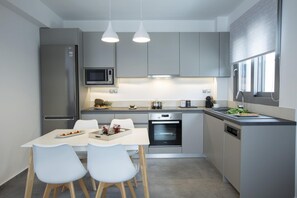 A modern kitchen with grey cabinets, a stainless steel refrigerator, a microwave, and a built-in oven. There is a dining table with white chairs and a tray of food. - Aloft Luxury - Villa Argia with sea view (Apolpena)