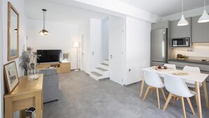 Una sala de estar moderna con un sofá gris, una mesa de centro de madera y un televisor de pantalla plana. Hay un comedor con una mesa y sillas blancas, y una cocina con gabinetes grises y un microondas.