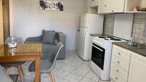 Eine kompakte Küche mit weißen Schränken, einem Herd und einem Kühlschrank. Es gibt eine Essecke mit Tisch und Stühlen, sowie ein graues Sofa.