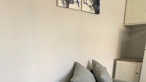 Ein graues Sofa mit zwei Kissen vor einer schlichten Wand, ein gefliestem Boden und einer Kommode mit einem gemusterten Deckel.