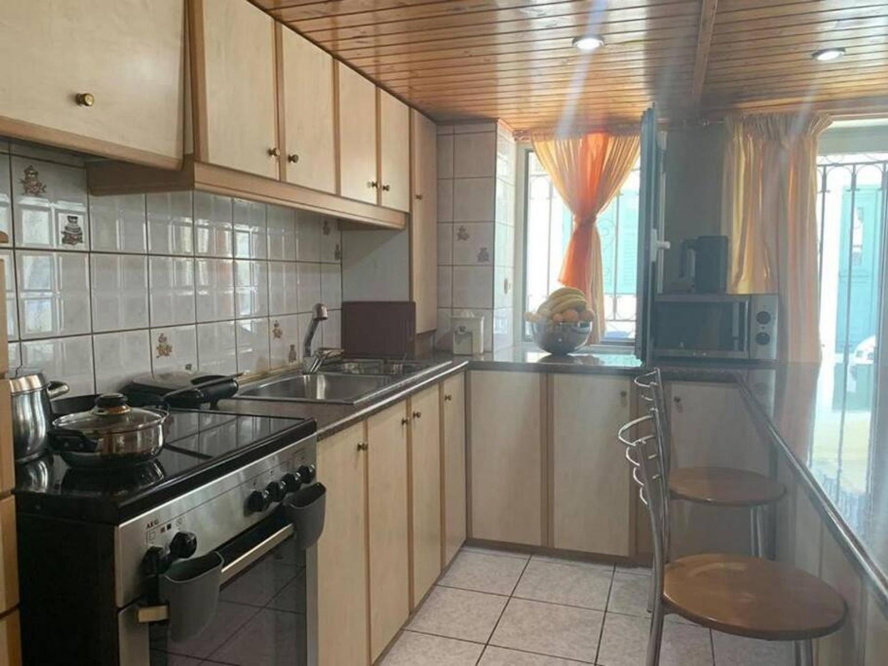 Una cocina con estufa, lavamanos y armarios. Se observa una ventana con cortinas y vistas al exterior.