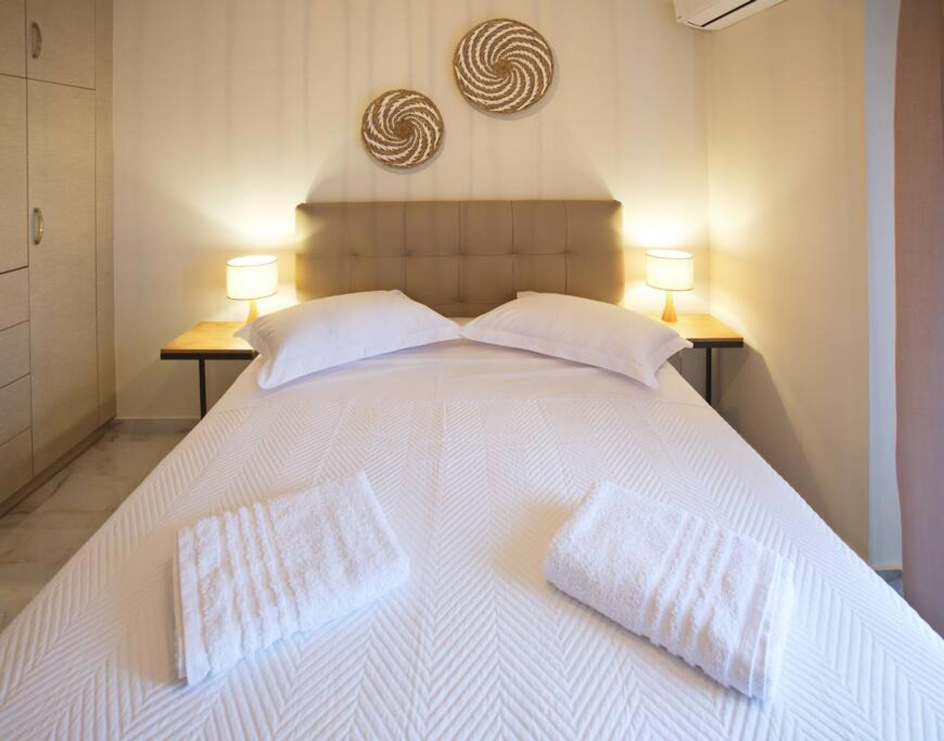 A neatly made bed with white linens and two pillows, flanked by two bedside tables with lamps, in a room with a wooden headboard and two wall-mounted decorative items.