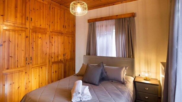 Une chambre avec un plafond en bois, un lit avec une literie grise, une table de chevet avec une lampe et une fenêtre avec des rideaux.