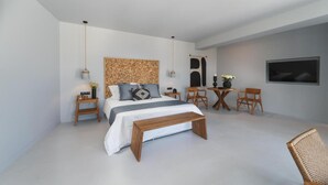 Een moderne slaapkamer met een groot bed, nachtkastjes, een houten bankje, een klein rond tafeltje met stoelen, en een tv aan de muur.