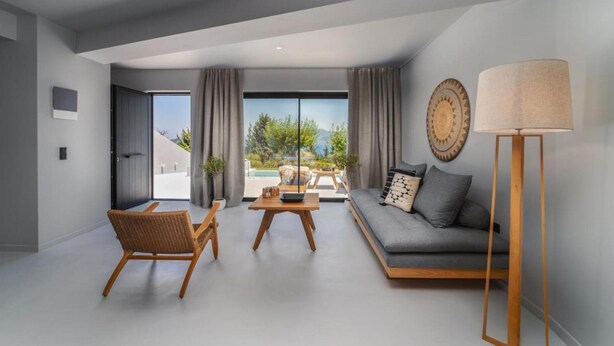 Een moderne woonkamer met een bank, een houten salontafel en een rieten fauteuil. Een groot raam biedt uitzicht op groen en een zwembad.