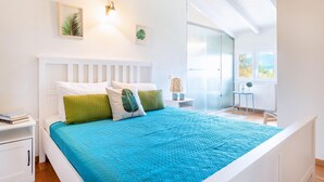 A modern bedroom with a white bed, blue bedding, and a view of the outdoors through a large window. - Villa Spyrelia in Dassia (Dassia)