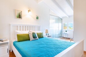 Ein modernes Schlafzimmer mit einem weißen Bett, blauer Bettwäsche und Blick ins Freie durch ein großes Fenster.