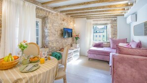 Una habitació acollidora amb una paret de pedra, bigues de fusta, una taula de dinar amb aliments per esmorzar, un sofà rosa i un televisor penjat a la paret.
