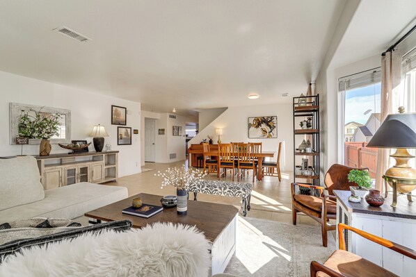 A spacious living area with a sofa, coffee table, and a dining table with chairs. There is a bookshelf with decorative items and a window with curtains. - 9 Mi to DIA: Group Getaway w/ Easy Denver Access (Commerce City)