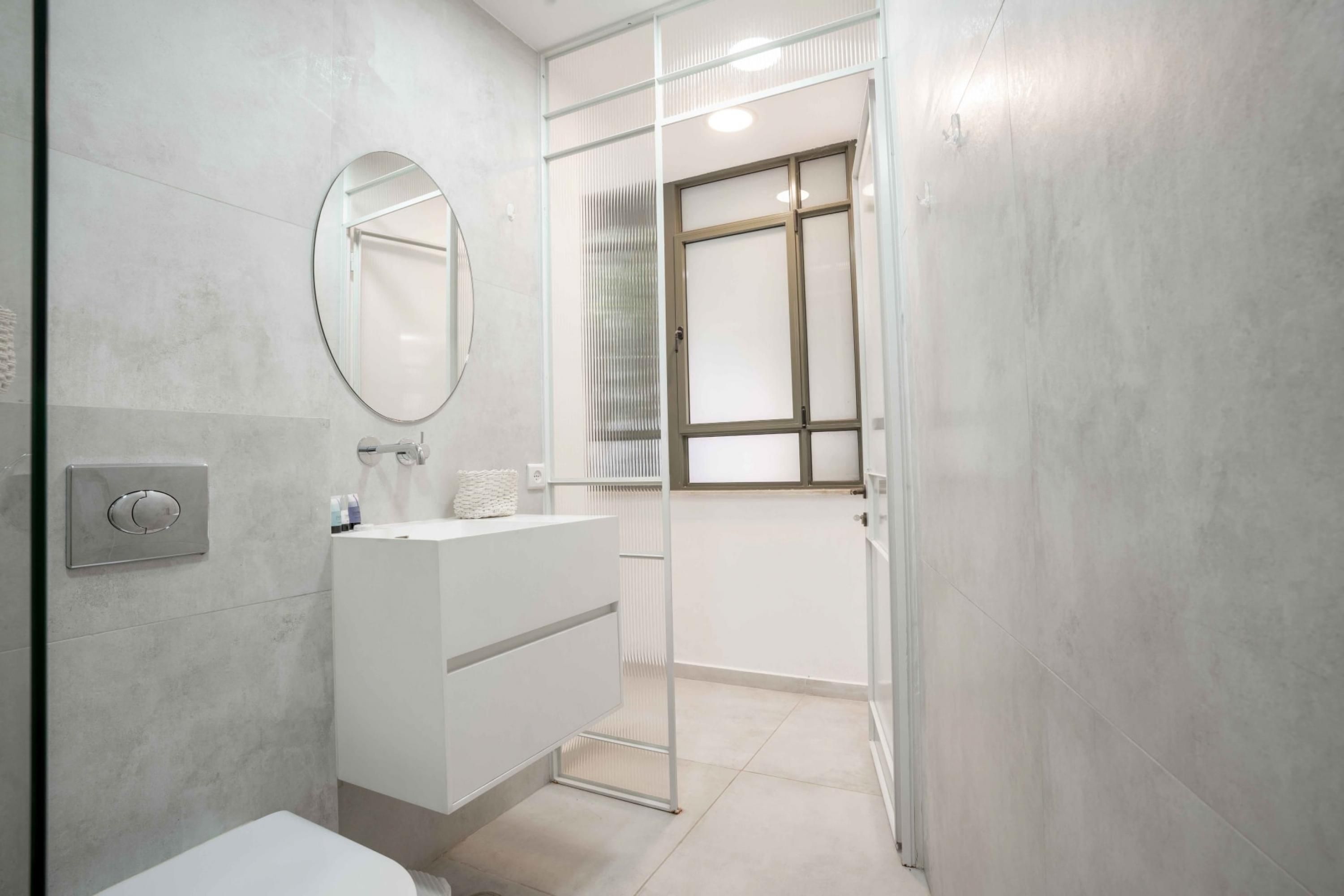 Une salle de bain moderne avec un meuble-lavabo blanc, un miroir rond et une porte en verre donnant accès à l’espace de la douche.