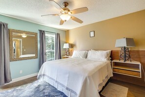 A bedroom with a bed, a ceiling fan, a nightstand with a lamp, a mirror, and a window with curtains.