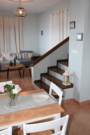 Un coin repas avec une table en bois, des chaises blanches et un vase de fleurs. Un escalier avec une rampe en bois mène à un autre niveau.