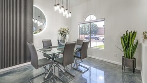 Un espace repas moderne avec une table en verre, des chaises en métal, un vase de fleurs et une grande fenêtre donnant sur l’extérieur.