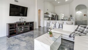 Un salon moderne doté d’un grand téléviseur à écran plat, d’une table basse en marbre, d’un canapé blanc et d’un coin cuisine avec des meubles blancs et des tabourets de bar.