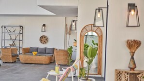 Ein stilvoller Lounge-Bereich mit Korbmöbeln, einem runden Couchtisch und einem kleinen Tisch mit einer Flasche. Übertöpfe und ein dekoratives Wandelement sind ebenfalls vorhanden.