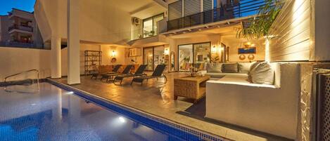 A modern outdoor swimming pool area with lounge chairs and a sofa, surrounded by a building with balconies and large windows.