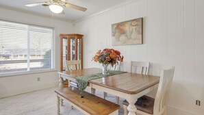 Een eetruimte met een houten tafel, bankjes en stoelen. Op de tafel staat een vaas met bloemen. Boven de tafel is een plafondventilator te zien.