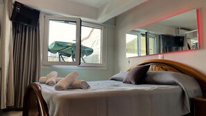 A hotel room with a bed, a window with curtains, a mirror, and a television mounted on the wall. - Rooftop Loft with Hydromassage (Nees Pagases)