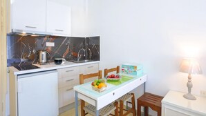 Et kompakt køkken med hvide skabe, en marmorbeklædt bagvæg, et lille spisebord med stole, og en skål med frugt på disken.