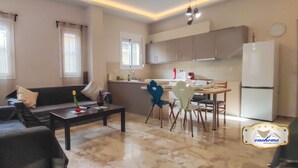 Ein Wohnzimmer mit Sofa, Couchtisch und Sesseln. Es gibt einen Essbereich mit Tisch und Stühlen. Die Küche verfügt über Schränke und einen Kühlschrank.