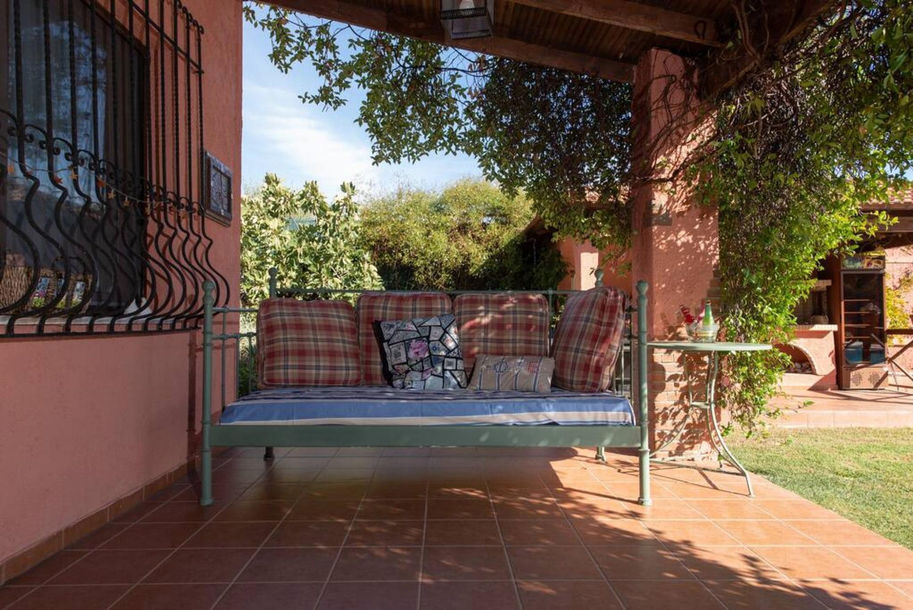 A covered outdoor seating area with a metal bench, a tiled floor, and a view of trees and a building in the background.