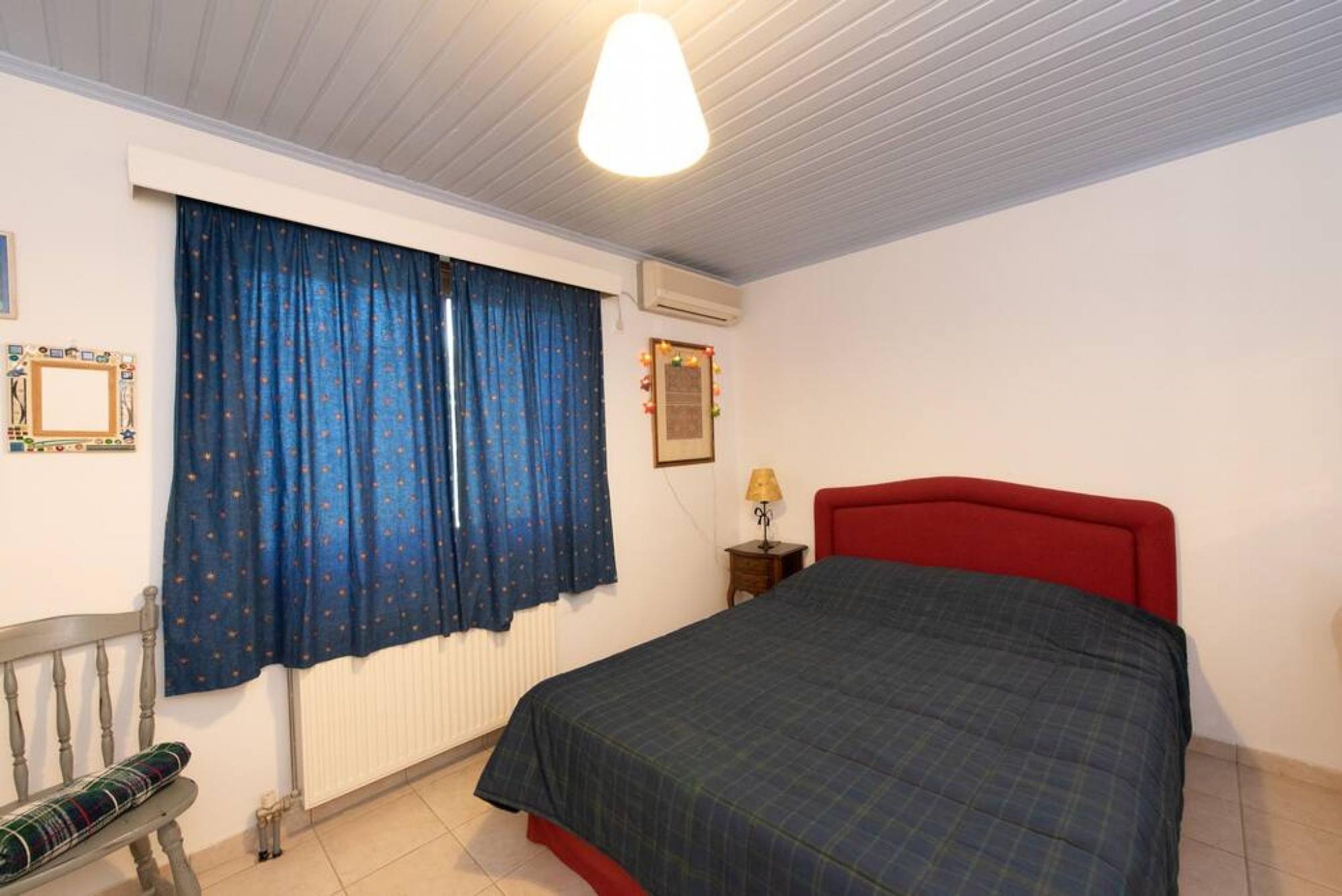 Un dormitorio con cama, cabecera roja, ventana con cortinas azules, mesita de noche con lámpara y radiador.