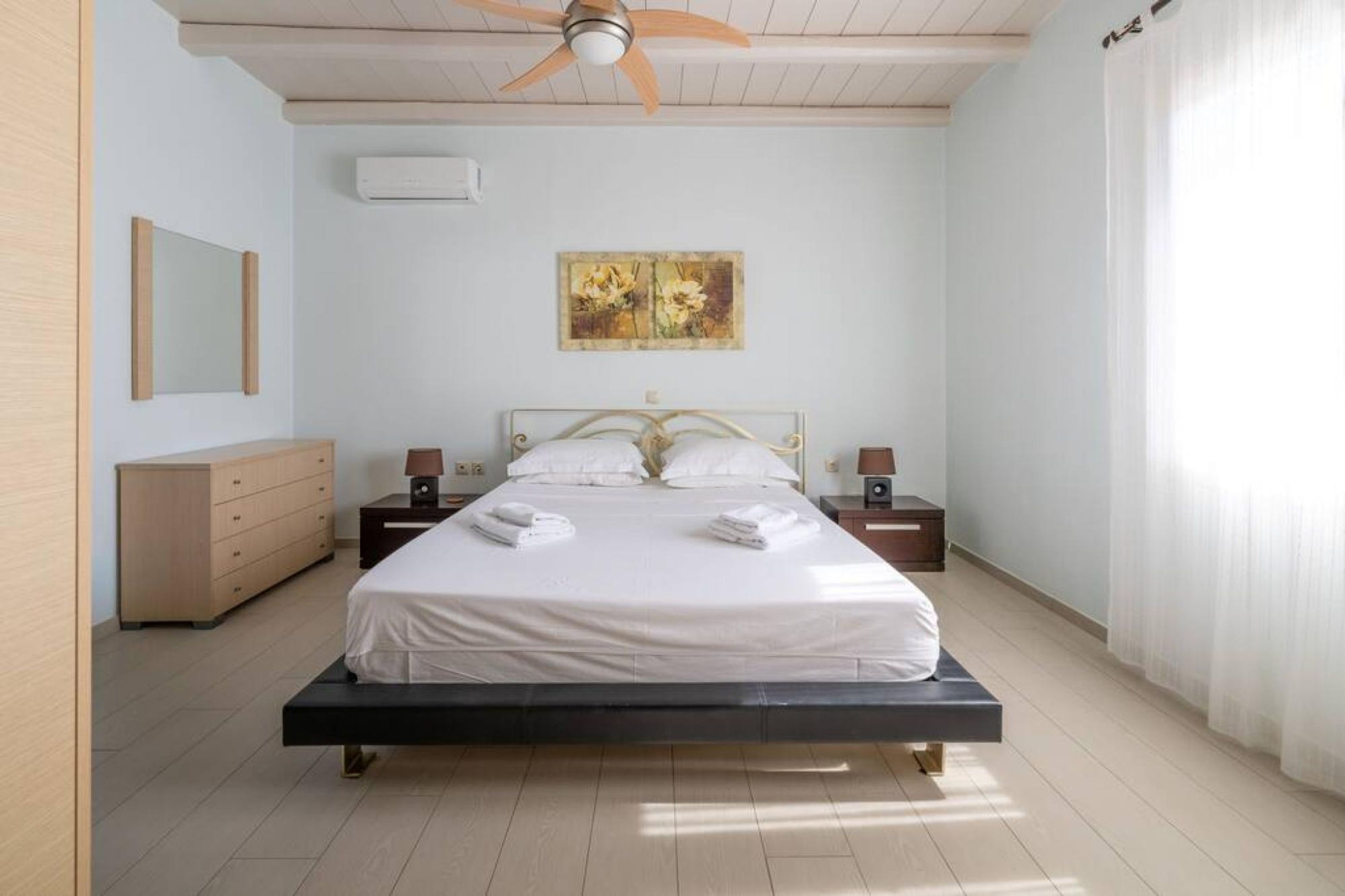 A neatly arranged bedroom with a bed, bedside tables, a ceiling fan, a painting, and a dresser.