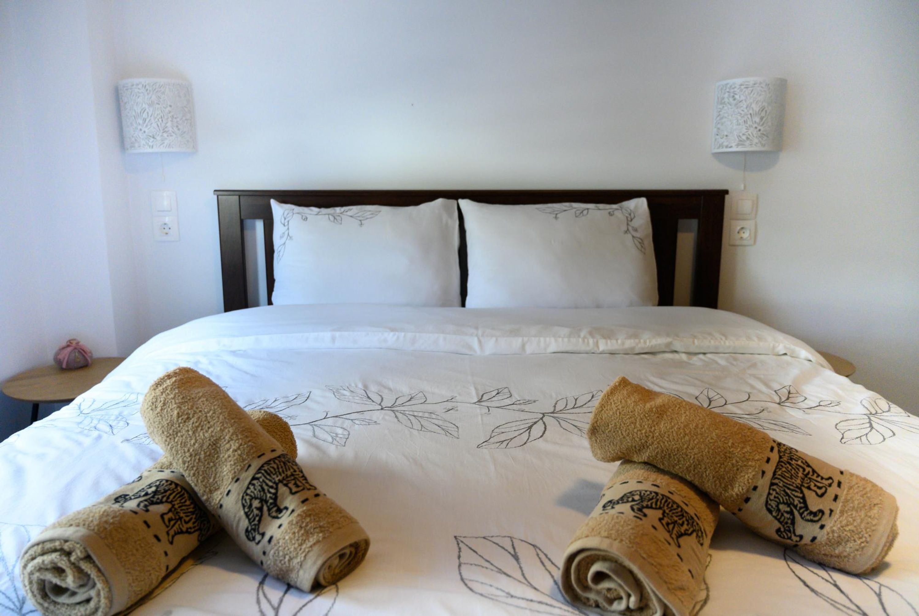 A neatly made bed with white linens and decorative pillows, two wall-mounted lamps, and a small round table with a pink object on it.