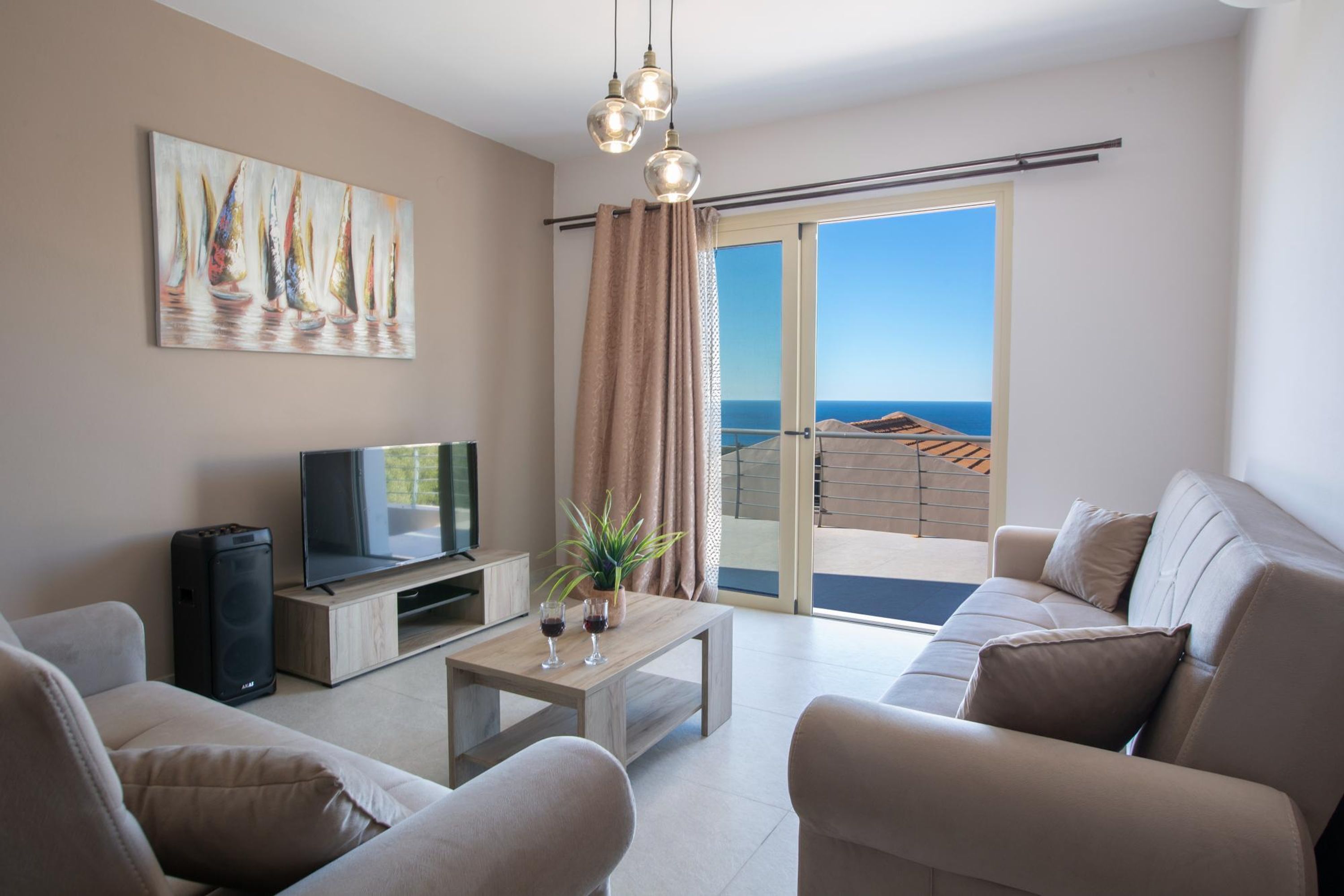 A modern living room with a flat-screen TV, a wooden coffee table, and beige sofas. A painting of sailboats is on the wall, and there's a balcony with a view of the sea.